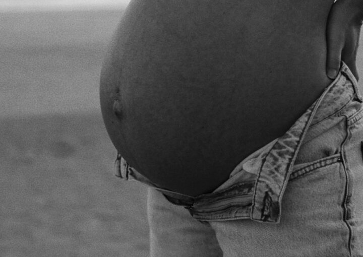 A silhouette shows a woman cradling her round pregnancy bump while standing on a sandy beach at sunset. Soft ocean waves and a warm golden sun create a tranquil atmosphere.